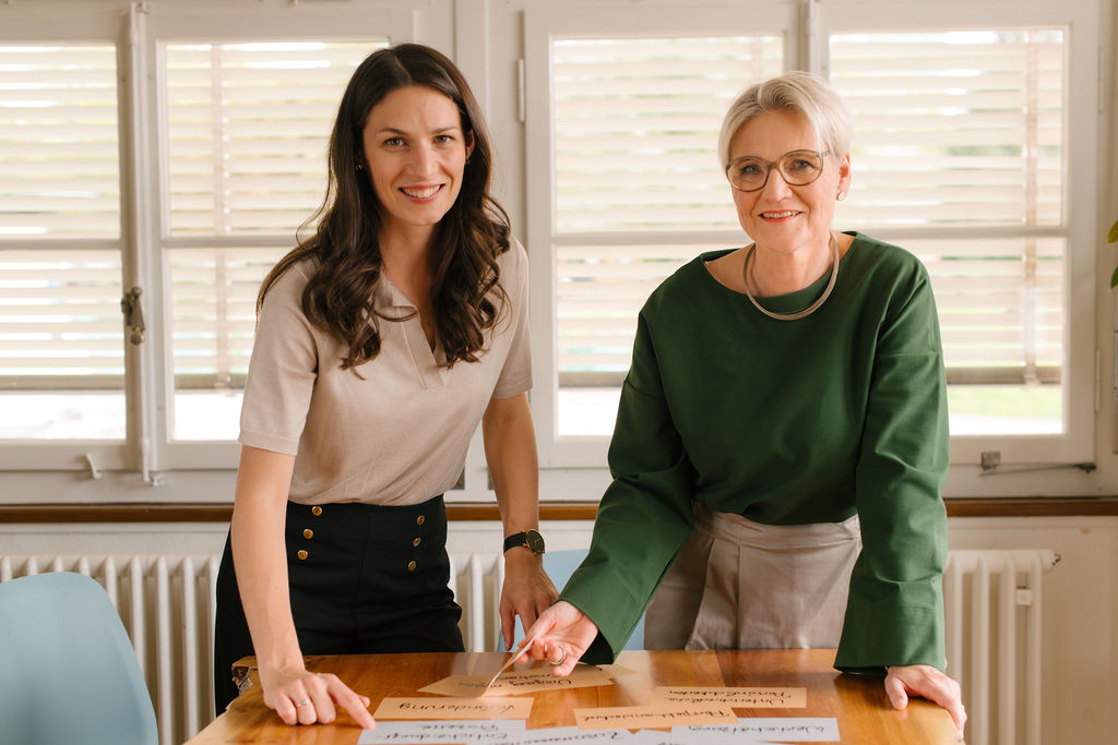 two women and mediator, Pamina Sulzberger and Susann Bongers are standing smiling at a table and looking at the camera in a light room